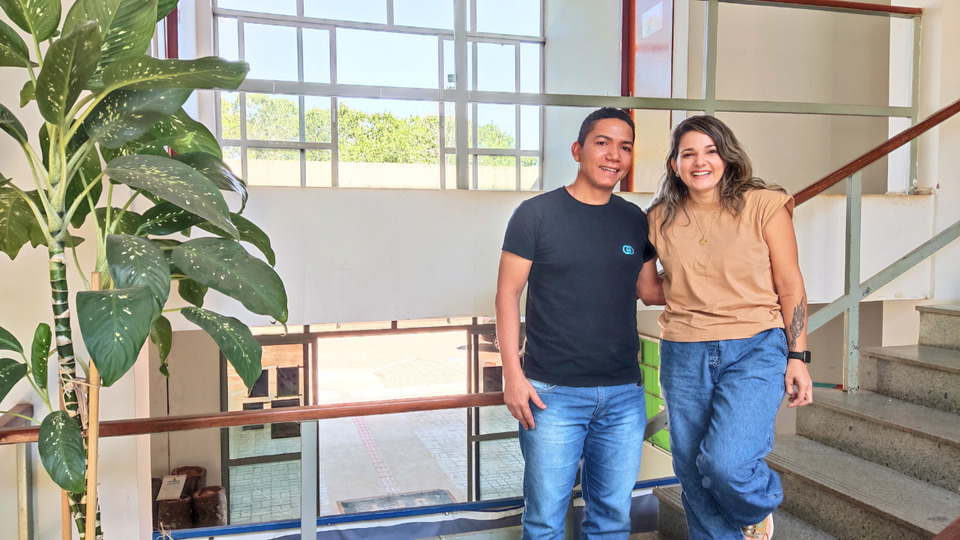 Foto colorida, com uma planta verde no canto esquerdo e duas pessoas pousando para uma foto em uma escada